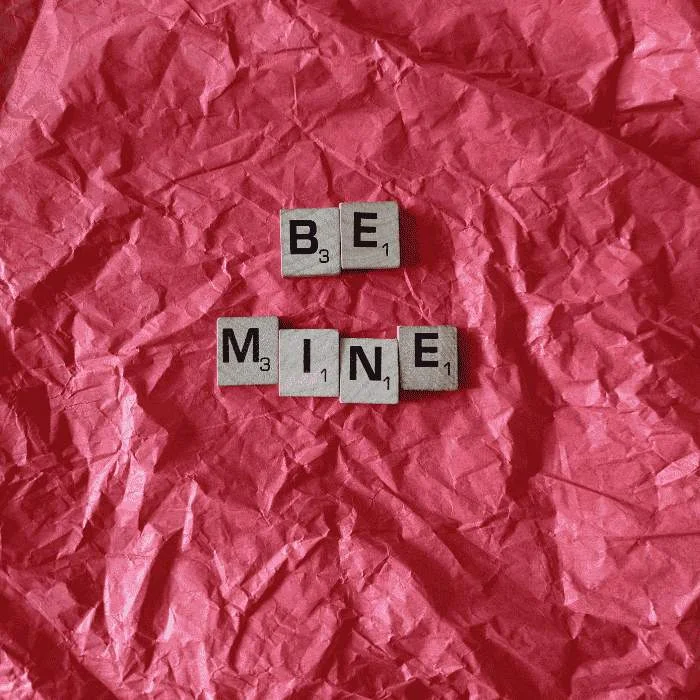 Ξύλινα γράμματα σχηματίζουν τη φράση “Be Mine” πάνω σε τσαλακωμένο κόκκινο φόντο.