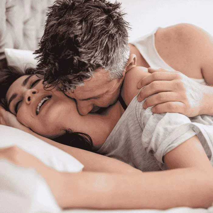 Άντρας και γυναίκα ξαπλωμένοι στο κρεβάτι αγκαλιάζονται σε τρυφερή στιγμή.