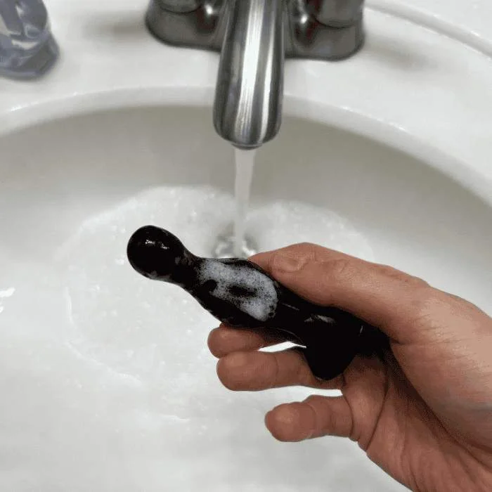 Χέρι πλένει μαύρο γυάλινο sex toy με σαπουνάδα κάτω από τρεχούμενο νερό σε λευκό νιπτήρα μπάνιου.
