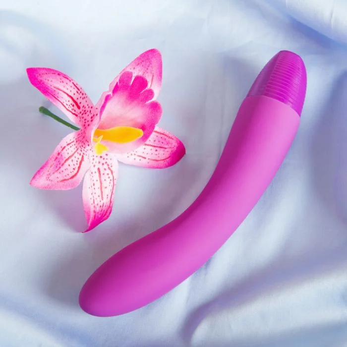 Ροζ G spot vibrator δίπλα σε ροζ λουλούδι, πάνω σε λευκό ύφασμα.