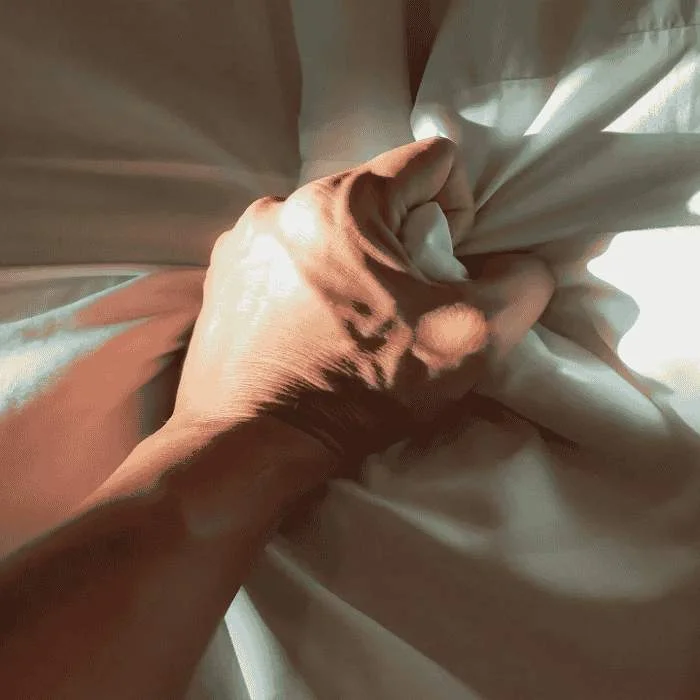 Ένα χέρι σφίγγει σφιχτά ένα λευκό ύφασμα, δημιουργώντας σκιές σε ζεστό φωτισμό. Γύρω από το χέρι, το ύφασμα είναι ελαφρώς τσαλακωμένο, υποδηλώνοντας ένταση σε ένα απαλά φωτισμένο περιβάλλον.
