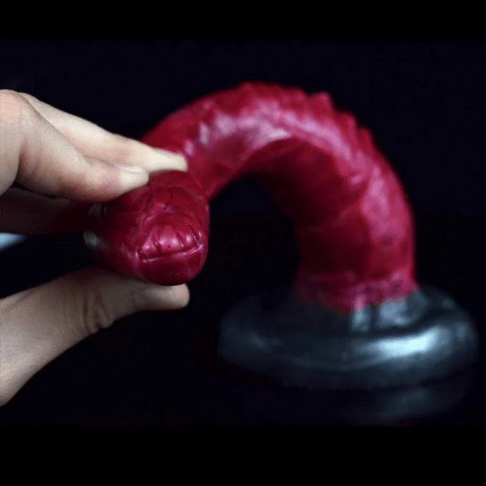 Χέρι πιέζει την εύκαμπτη κεφαλή ενός dildo σε σχήμα φιδιού, σε βαθύ μπορντό χρώμα με λεπτομερή υφή λέπιας και καμπυλωτό κορμό. Φόντο σκοτεινό, τονίζει το αντικείμενο.