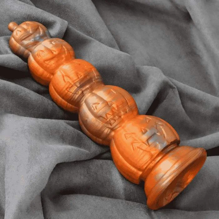 Fantasy dildo σε πορτοκαλί απόχρωση με ανάγλυφα σχέδια jack-o’-lantern σε σειρά, τοποθετημένο επάνω σε μαλακό γκρι ύφασμα.