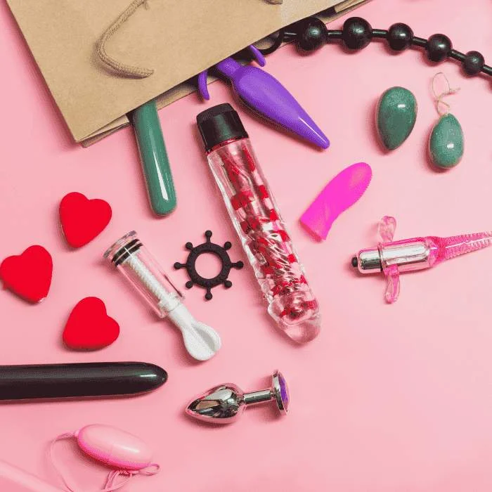 Ανοιχτή χάρτινη σακούλα με sex toys που έχουν πέσει πάνω σε ροζ επιφάνεια.