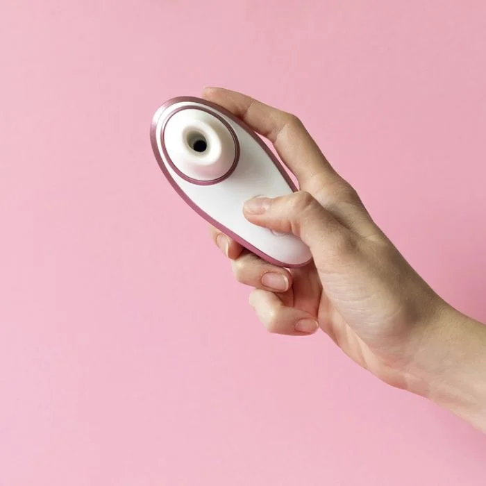Χέρι κρατά λευκό satisfyer με ροζ λεπτομέρειες πάνω σε ροζ φόντο.
