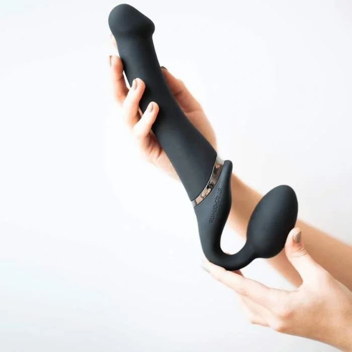 Γυναικεία χέρια κρατούν μαύρο strapless strap-on από σιλικόνη, με εργονομική καμπύλη και διπλή άκρη, ιδανικό για hands-free διείσδυση.