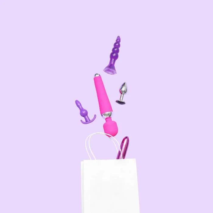 Διάφορα ροζ και μωβ sex toys βγαίνουν από λευκή χάρτινη σακούλα σε μωβ φόντο.