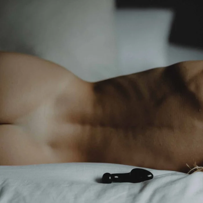 Η πλάτη ενός ατόμου βρίσκεται οριζόντια σε ένα κρεβάτι, με ορατούς μύες. Ένα μικρό μαύρο αντικείμενο είναι τοποθετημένο κοντά στο λευκό σεντόνι, υπό απαλό, διάχυτο φωτισμό.