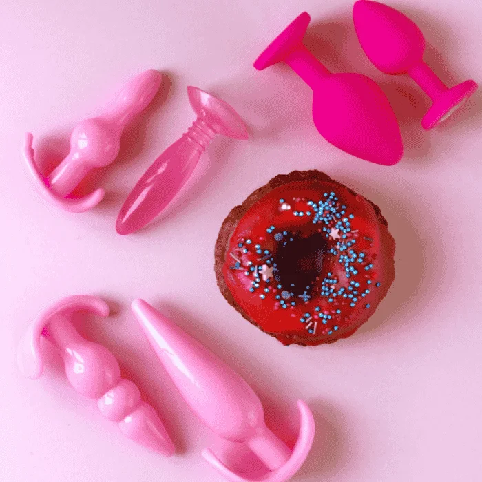 Διάφορα πολύχρωμα βύσματα σιλικόνης είναι τοποθετημένα γύρω από ένα σοκολατένιο ντόνατ με κόκκινο γλάσο και μπλε τρούφα, πάνω σε μια ροζ επιφάνεια.