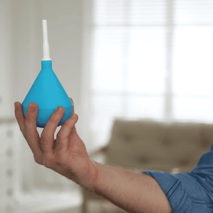 Ανδρικό χέρι κρατά ένα μπλε πρωκτικό κλύσμα με λευκό ακροφύσιο, μπροστά από φωτεινό φόντο σαλονιού με θολό παράθυρο στο βάθος.