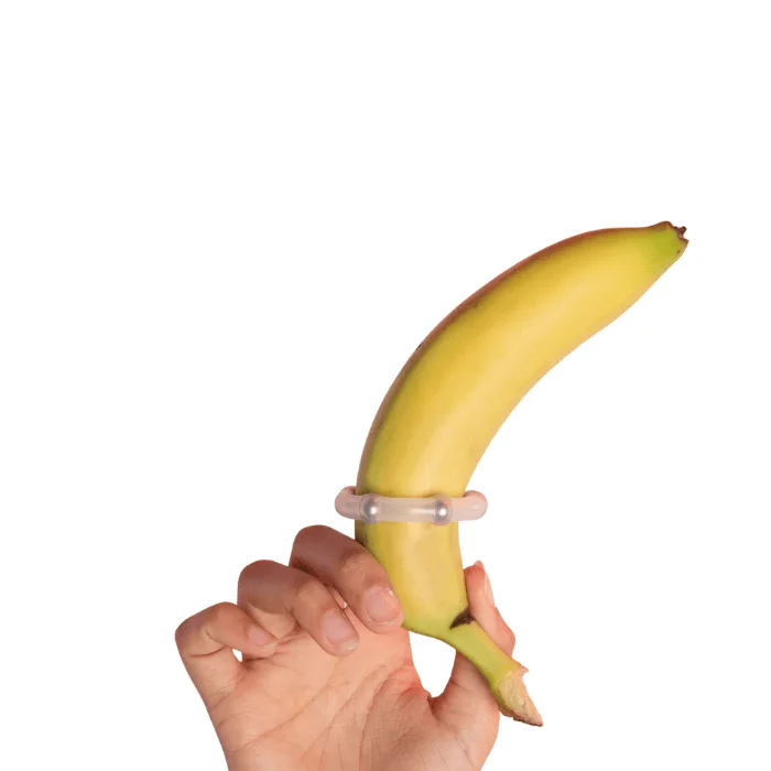 Χέρι κρατάει μια μπανάνα που φοράει διάφανο δαχτυλίδι πέους με χάντρες, σε λευκό φόντο.