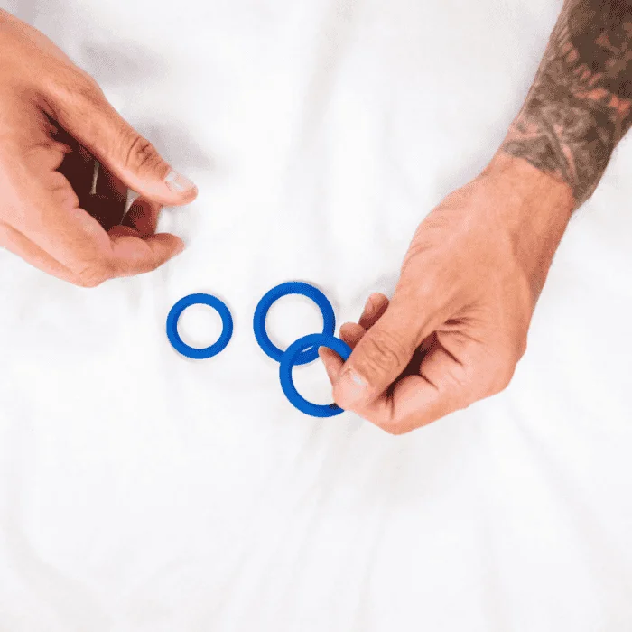 Ανδρικά χέρια με τατουάζ κρατούν τρία μπλε δαχτυλίδια πέους διαφορετικών μεγεθών, με λεία επιφάνεια, πάνω σε λευκό σεντόνι.