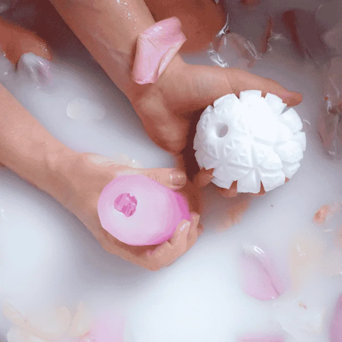 Χέρια που κρατούν ένα ανδρικό αυνανιστήρι και έναν κλειτοριδικό αναρροφητή μέσα σε γαλακτερό νερό μπάνιου, διακοσμημένο με ροδοπέταλα.