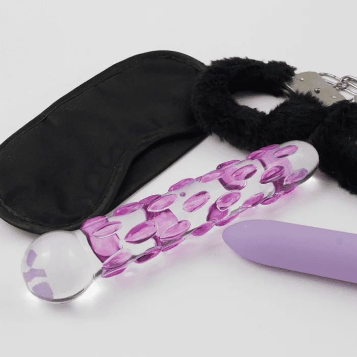 Διάφορα sex toys και αξεσουάρ όπως γυάλινο dildo, μωβ δονητής, μάσκα ματιών και χειροπέδες σε ουδέτερη επιφάνεια.