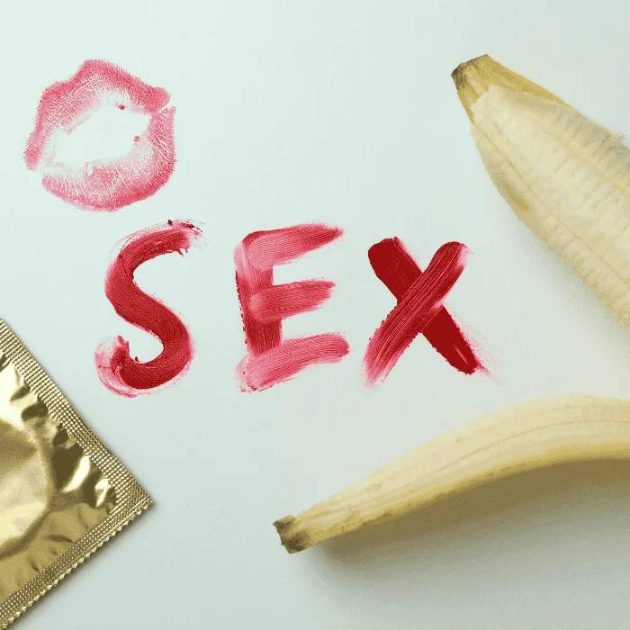 Προφυλακτικό και ανοιγμένη μπανάνα δίπλα στη λέξη sex και αποτύπωμα χειλιών απο κραγιόν σε λευκή επιφάνεια.