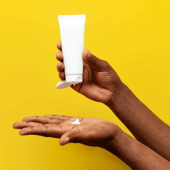 Χέρι που κρατά λευκό σωληνάριο και ρίχνει κρέμα ή gel στην παλάμη, σε κίτρινο φόντο.