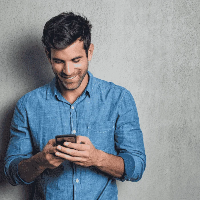 Άντρας με μπλε πουκάμισο κοιτά το κινητό του και χαμογελά σε γκρι φόντο.
