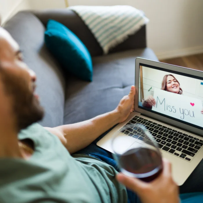 Άντρας κάθεται σε καναπέ και βλέπει βιντεοκλήση σε laptop κρατώντας ποτήρι κρασί.