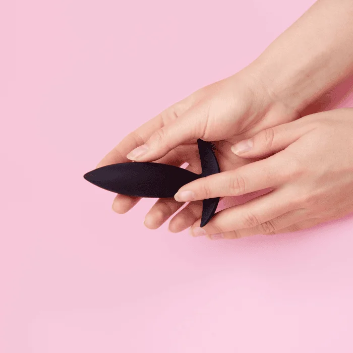 Γυναικεία χέρια κρατούν μαύρη πρωκτική σφήνα σιλικόνης πάνω σε ροζ φόντο.