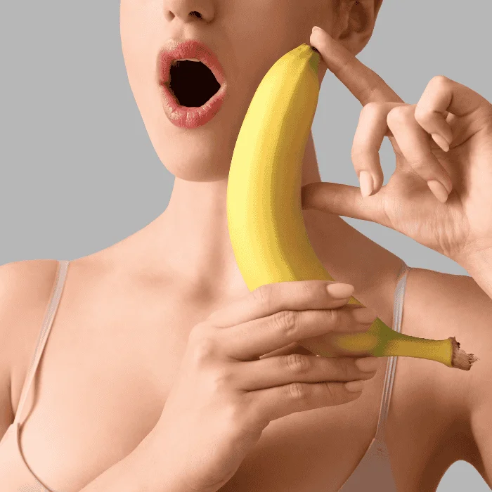 Γυναίκα με ανοιχτό στόμα κρατά μπανάνα μπροστά στο πρόσωπο, σε ουδέτερο φόντο.