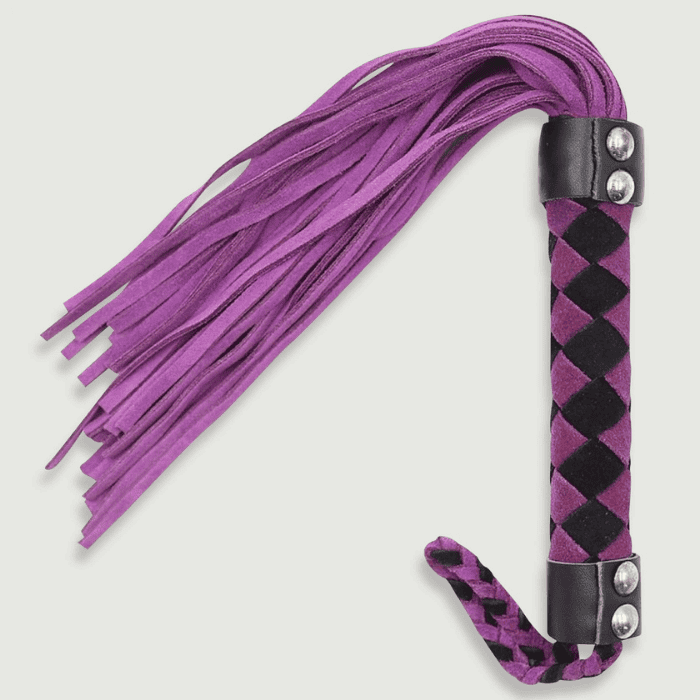 Ένα μοβ σουέτ μαστίγιο με μαύρη λαβή με καρό σχέδιο και ασημένια διακοσμητικά καρφιά βρίσκεται σε ένα απλό λευκό φόντο. Οι λωρίδες του είναι μακριές και ρέουν χαλαρά.