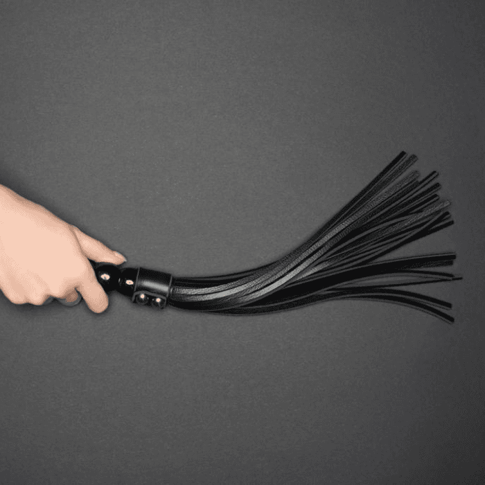 Ένα χέρι κρατά ένα μαύρο δερμάτινο μαστίγιο με πολλές λωρίδες να εκτείνονται προς τα έξω, σε ένα απλό γκρι φόντο.