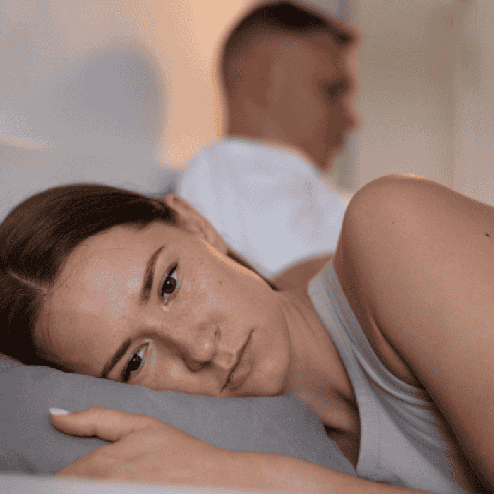 Γυναίκα ξαπλωμένη στο πλάι κοιτάζει μπροστά, ενώ ο άνδρας κάθεται στο πίσω μέρος του κρεβατιού γυρισμένος προς την άλλη πλευρά.