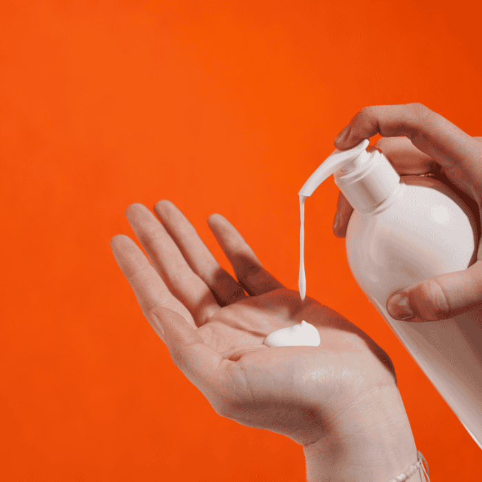 Χέρι αντλεί λευκή λοσιόν από μπουκάλι σε ανοιχτή παλάμη, πορτοκαλί φόντο.