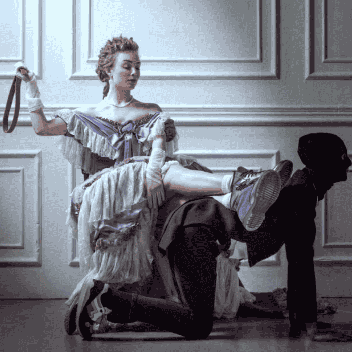 Γυναίκα με περίτεχνο φόρεμα εποχής κρατά ζώνη και ακουμπά τα πόδια της στην πλάτη μασκοφόρου άνδρα στα τέσσερα.