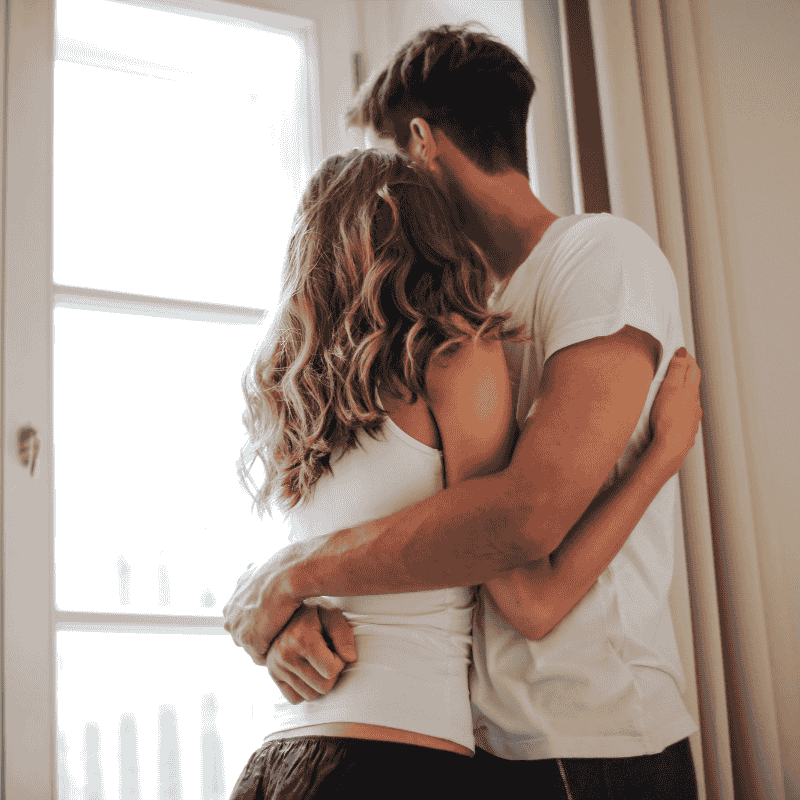Αγκαλιασμένο Ζευγάρι με Λευκά Μπλουζάκια Μπροστά σε Παράθυρο Νεαρό ζευγάρι αγκαλιάζεται μπροστά σε παράθυρο με φυσικό φως, φορώντας απλά ρούχα και εκπέμποντας οικειότητα και τρυφερότητα.